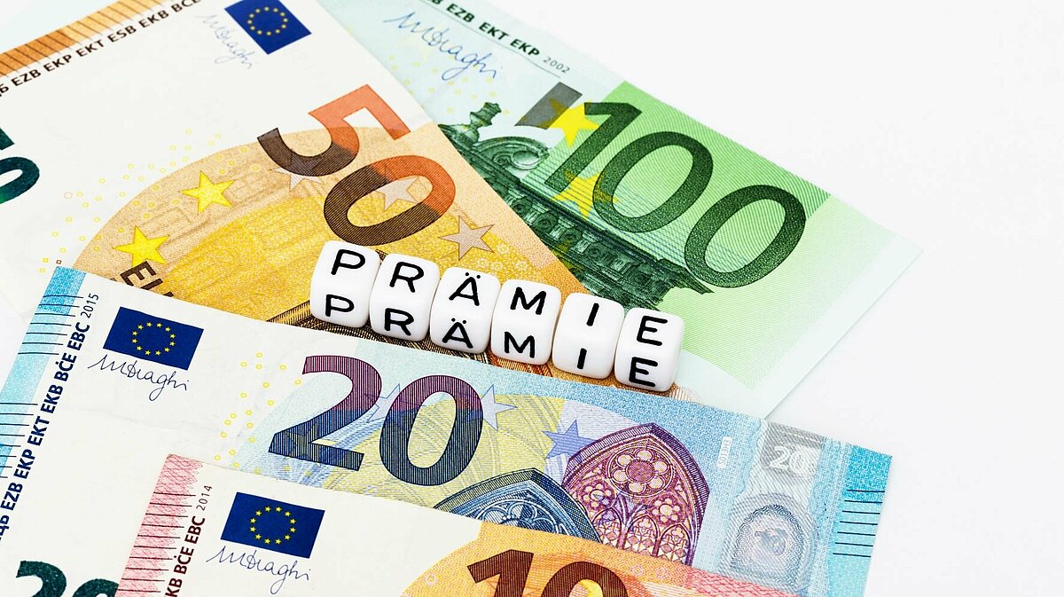 Mehrer Euro-Scheine liegen auf dem Tisch. Darauf platziert ein paar Würfel, die zusammengestellt das Wort "Prämie" ergeben.