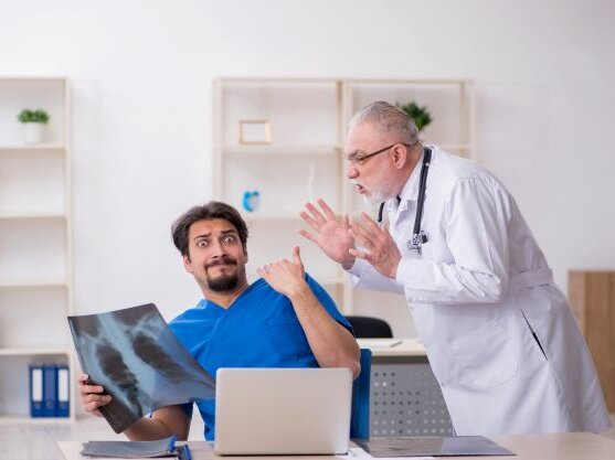 Junger Arzt sitzt mit einem Röntgenbild in der Hand am Schreibtisch und reißt erschrocken die Augen auf. Von der rechten Seite brüllt ihn ein älterer Arzt mit erhobenen Händen an.