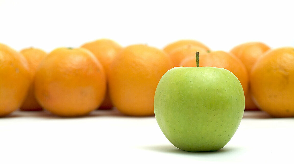 Ein grüner Apfel liegt im Vordergund. Dahinter viele Orangen.